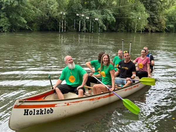 Eine Mannschaft in grünen ZITRONENSTOLZ- und schwarzen SÜDKOLA-Shirts lacht im Boot »Holzklob«, klatschnass vom Regen